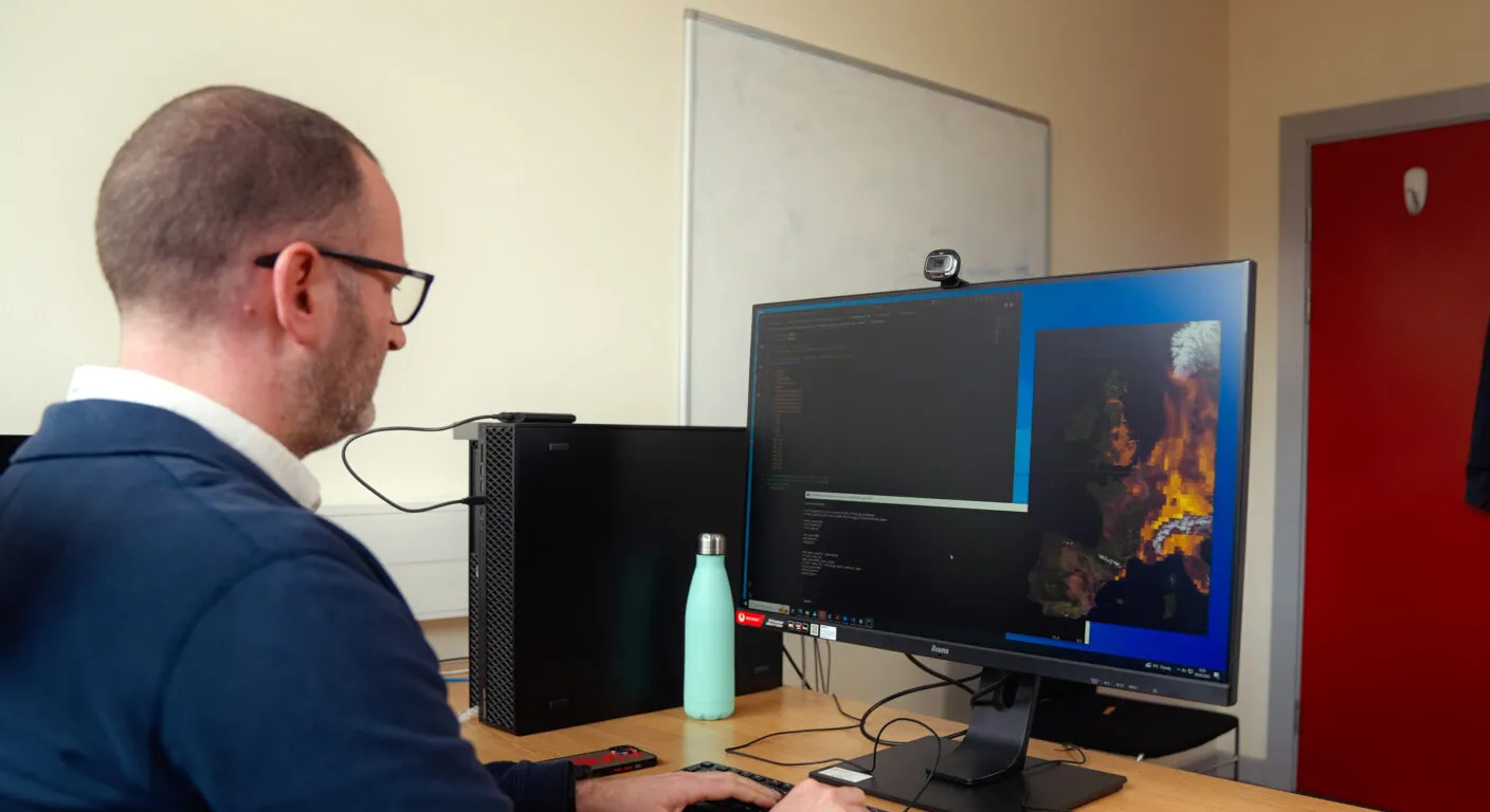 A man in a blue shirt works at a desk with a computer displaying colorful data visualizations on its screen.