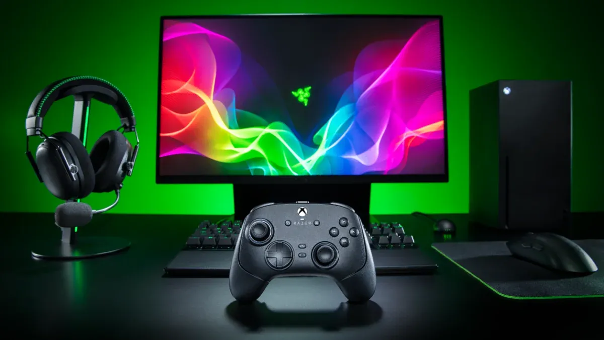 A black Razer Wolverine V4 controller sits on a desk with a monitor, headset, keyboard, mouse, and PC tower under green lighting.