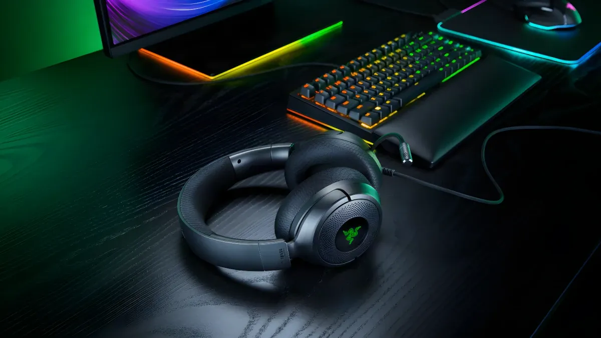 Dark gray Razer Kraken V4 X headphones with green logo rest on a wooden desk beside RGB-lit keyboard and monitor.