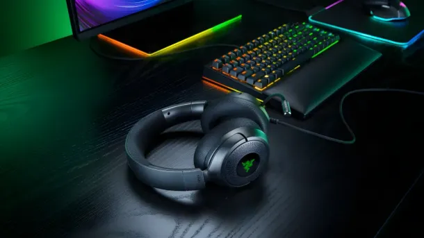 Dark gray Razer Kraken V4 X headphones with green logo rest on a wooden desk beside RGB-lit keyboard and monitor.
