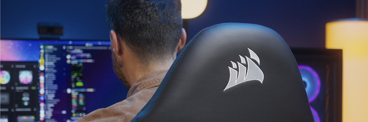 A person sits in a black Corsair gaming chair with the brand logo on the backrest, facing a computer monitor in a dimly lit room with ambient lighting.