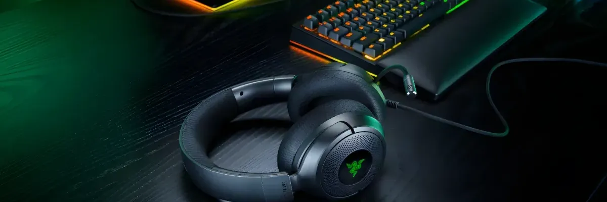 Dark gray Razer Kraken V4 X headphones with green logo rest on a wooden desk beside RGB-lit keyboard and monitor.