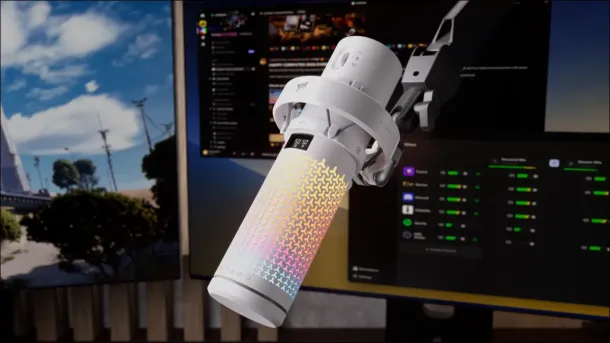 A white Corsair VOXARA microphone with colorful RGB lighting is mounted on a desk arm in front of dual monitors displaying gaming and audio software.