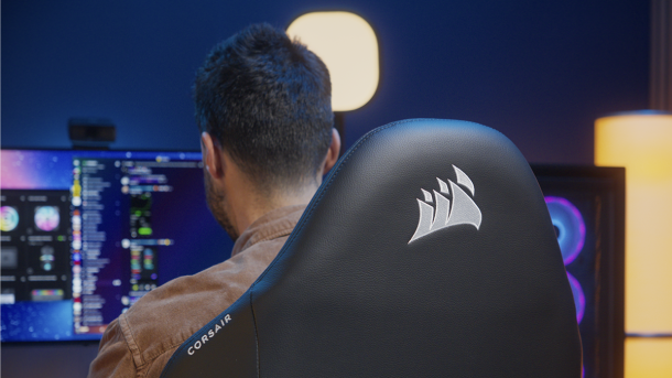 A person sits in a black Corsair gaming chair with the brand logo on the backrest, facing a computer monitor in a dimly lit room with ambient lighting.